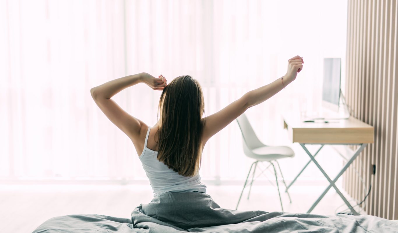 Back view of young woman stretching on unmade bed after waking up and looking at city view in the window. Motivation concept